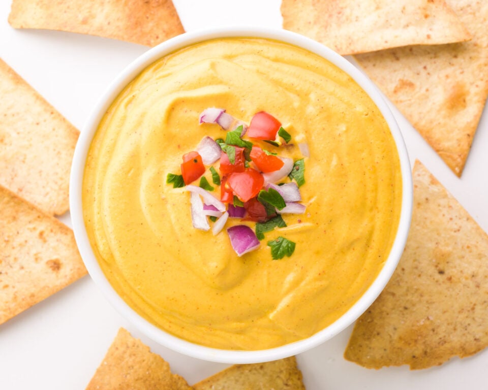 Looking down on a bowl of vegan nacho cheese with chopped veggies on top and tortilla chips all around the bowl.