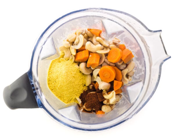 Looking down on a blender jar with ingredients like carrots, cashews, nutritional yeast flakes, chili powder, and more.