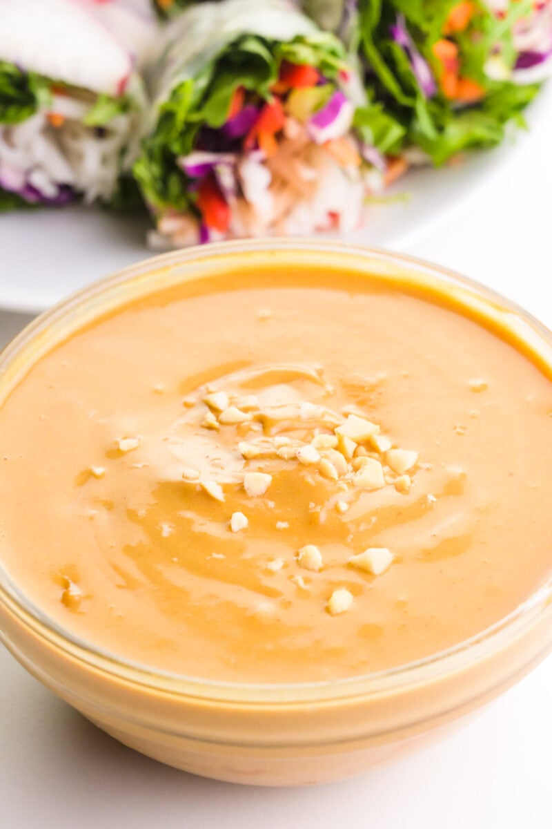 A bowl of vegan peanut sauce has chopped peanuts on top. There's a plate of veggie spring rolls in the background.