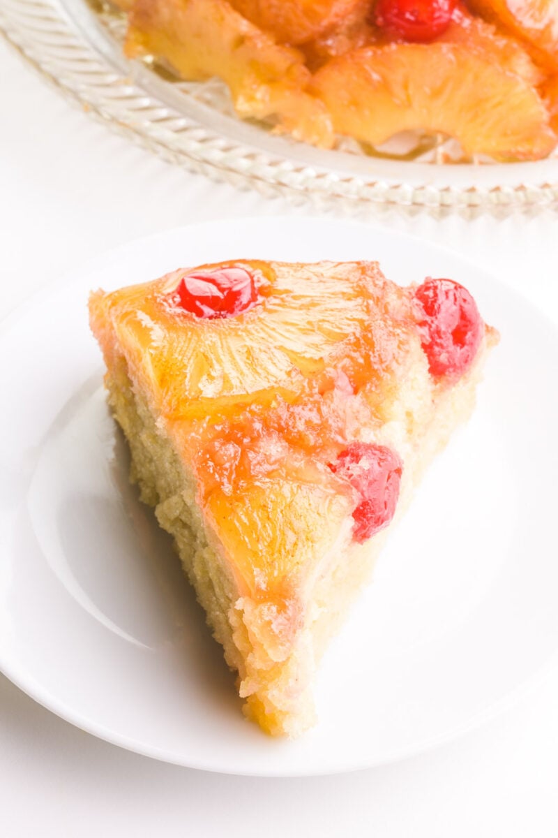 A slice of vegan pineapple upside down cake shows pineapples and maraschino cherries on top. The rest of the cake is behind it.