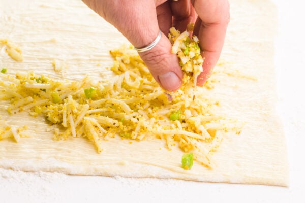 A hand is distributing a cheese mixture over buttered pastry sheet.