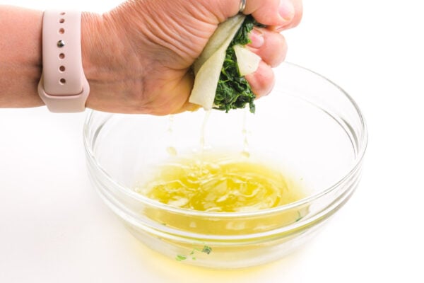A hand holds frozen spinach wrapped in a paper towel and is squeezing liquid from the spinach out into a bowl.