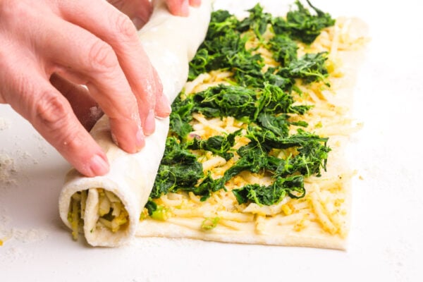 Hands are rolling up pastry with a spinach and cheese filling.