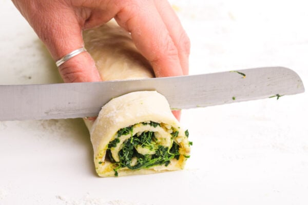 A hand holds a serrated knife, using it to cut slices out of a spinach pastry log.