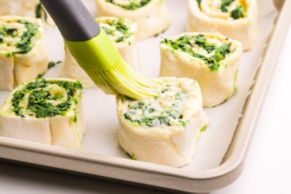 A pastry brush is brushing melted butter over pastry rolls on a baking sheet lined with parchment paper.