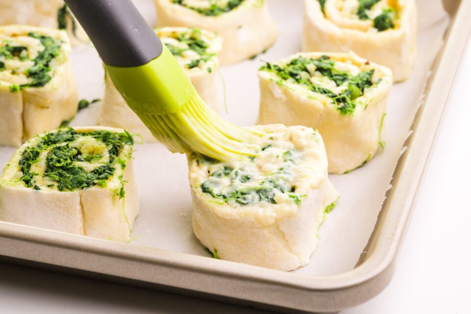 A pastry brush is brushing melted butter over pastry rolls on a baking sheet lined with parchment paper.