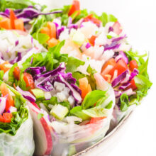 A bowl holds several vegan spring rolls, showing colorful vegetables in each roll. There's a bowl of peanut dipping sauce in the background.