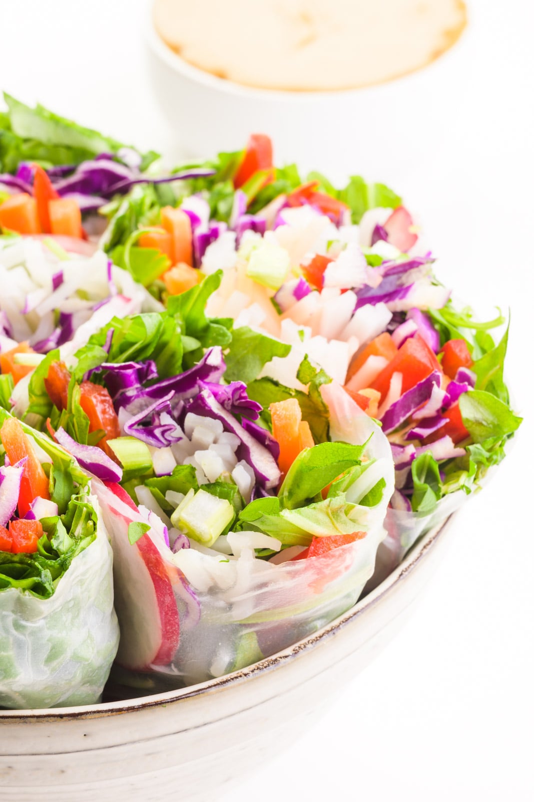 A bowl holds several vegan spring rolls, showing colorful vegetables in each roll. There's a bowl of peanut dipping sauce in the background.