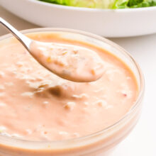 A spon full of vegan thousand island dressing hovers over a bowl full of the dressing. It sits in front of a bowl of salad, barely visible in the background.