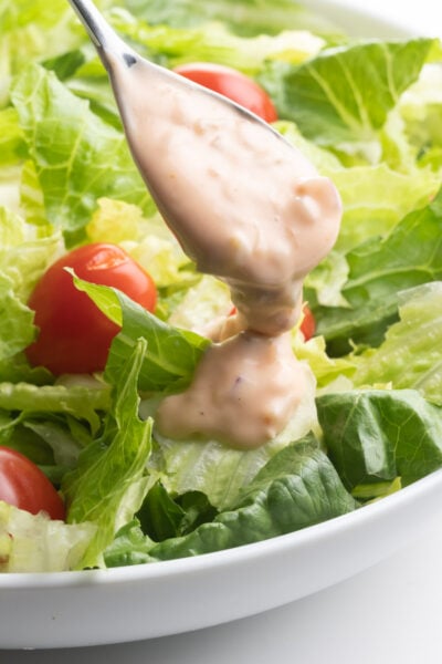 A spoon drizzles vegan thousand island dressing over a bowl of crispy greens and cherry tomatoes.