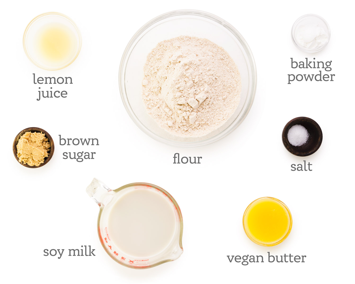 Ingredients are laid out on a white table. The labels next to each ingredient reads, flour, baking powder, salt, vegan butter, vegan milk, brown sugar, and lemon juice.