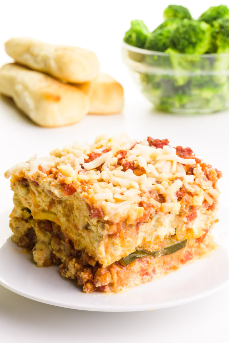 A slice of zucchini lasagna sits on a plate. There are breadsticks in the background and a bowl of steamed broccoli.