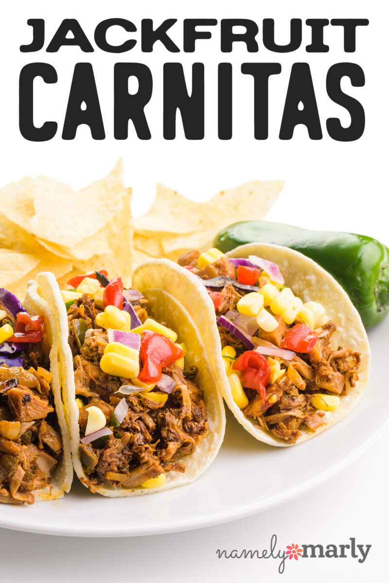 Three jackfruit carnitas tacos topped with corn, tomatoes, and red onion, accompanied by tortilla chips and a jalapeño.