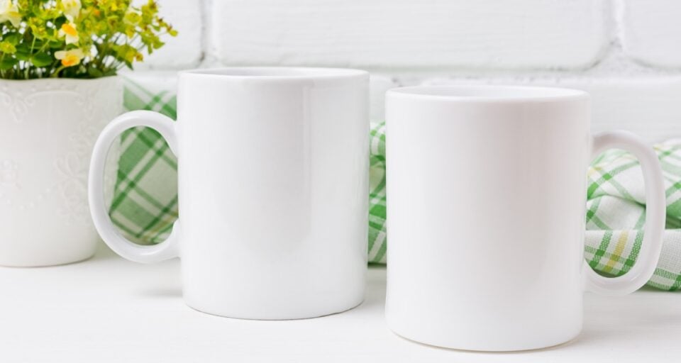 Two coffee mugs sit on a counter . There's a kitchen towel behind them a plant beside them.