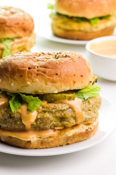 A fried tofu sandwich sits on a plate in front of other plated sandwiches. There's a bowl with mayo sauce in the middle.