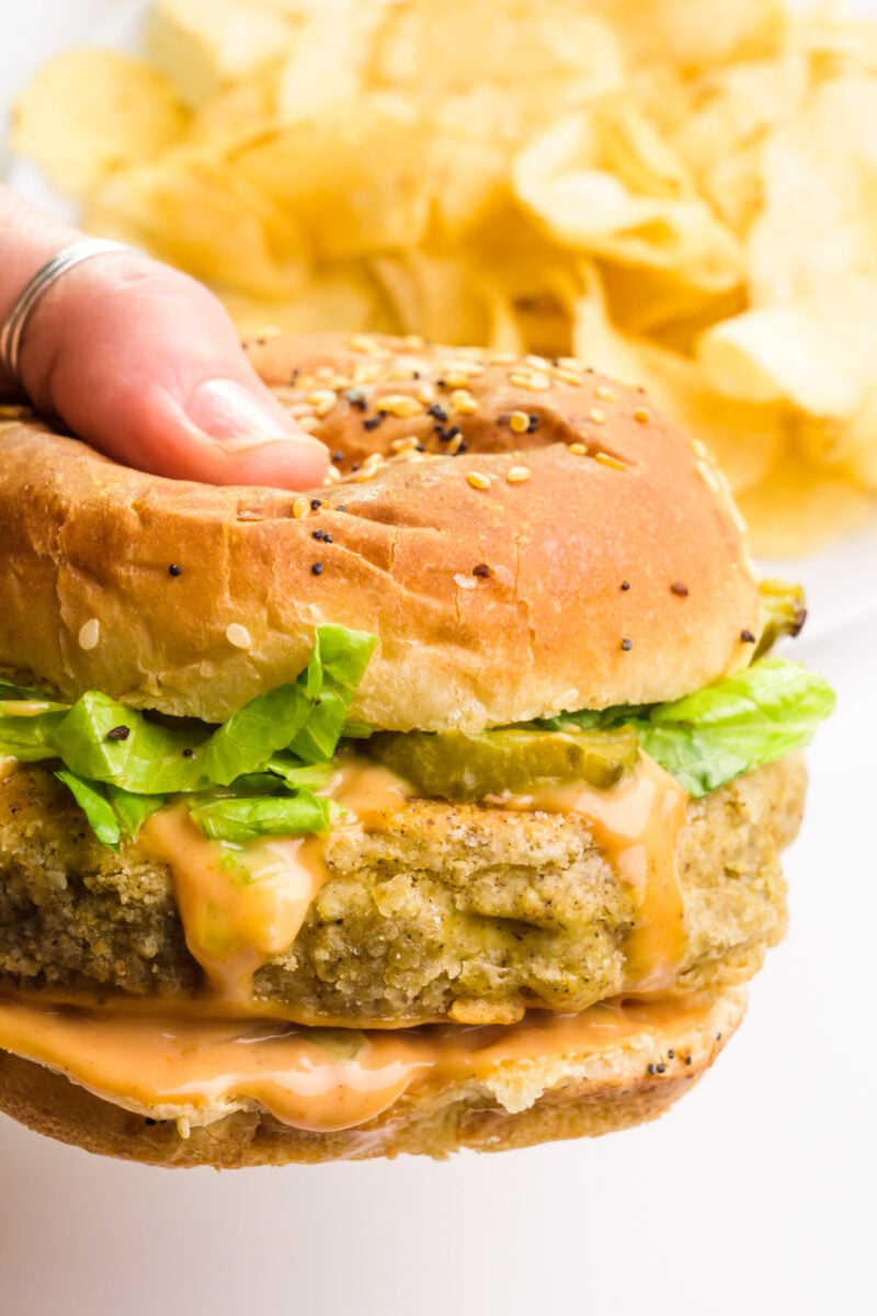 A hand holds a crispy tofu sandwich . There's a plate with potato chips in the background.