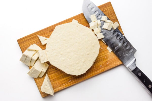 Tofu is on a cutting board sitting next to pieces that have been cut off with a knife.