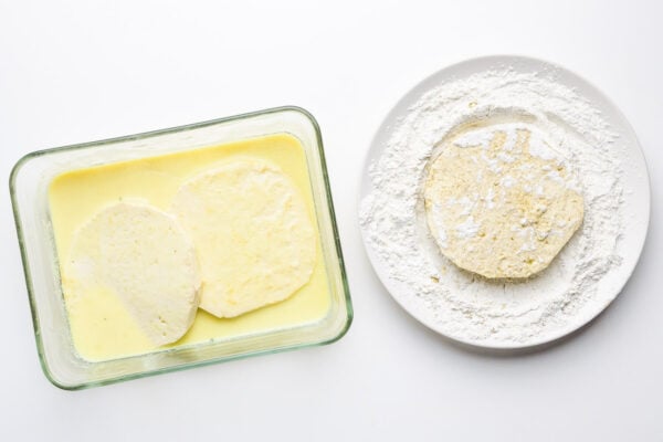 A rectangle baking dish has a soy milk mixture in it and two tofu patties. A plate sits next to it with a flour mixture and another tofu patty in that.