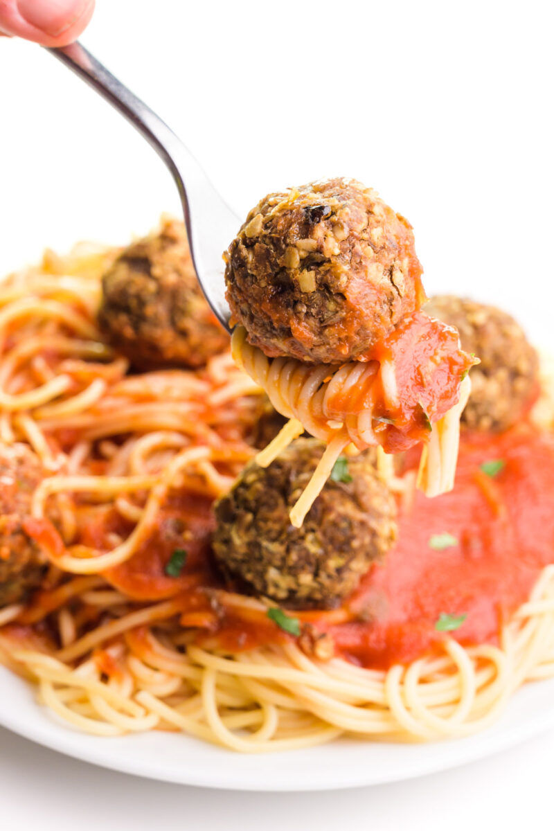 A fork holds a lentil meatball on top of spaghetti and red sauce. It hovers over a plate of more of the dish.