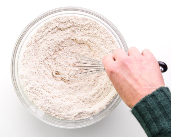 A hand holds a whisk, stirring a flour mix in a bowl.