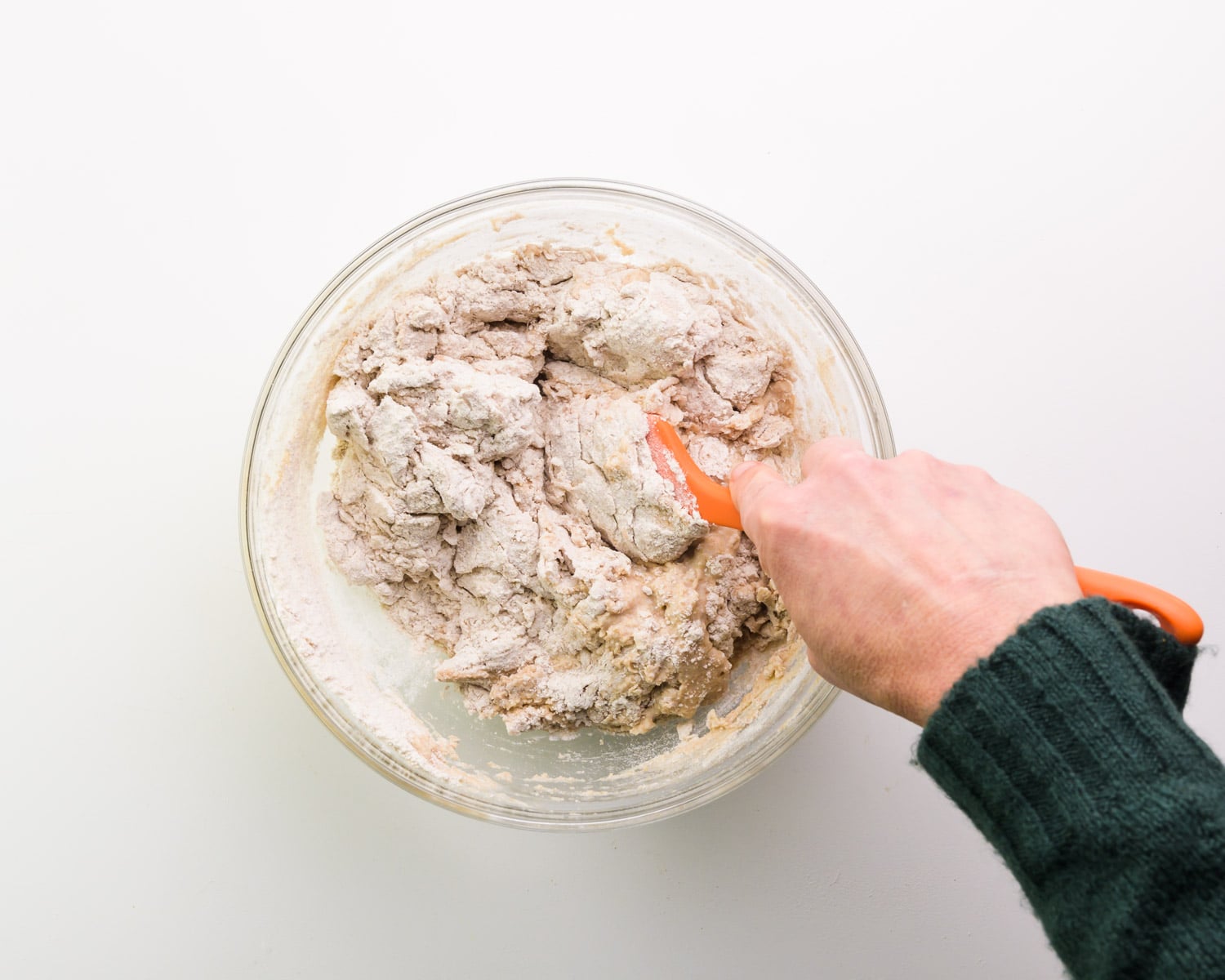 A hand holds an orange spatula, using it to stir a flour mixture in a bowl.