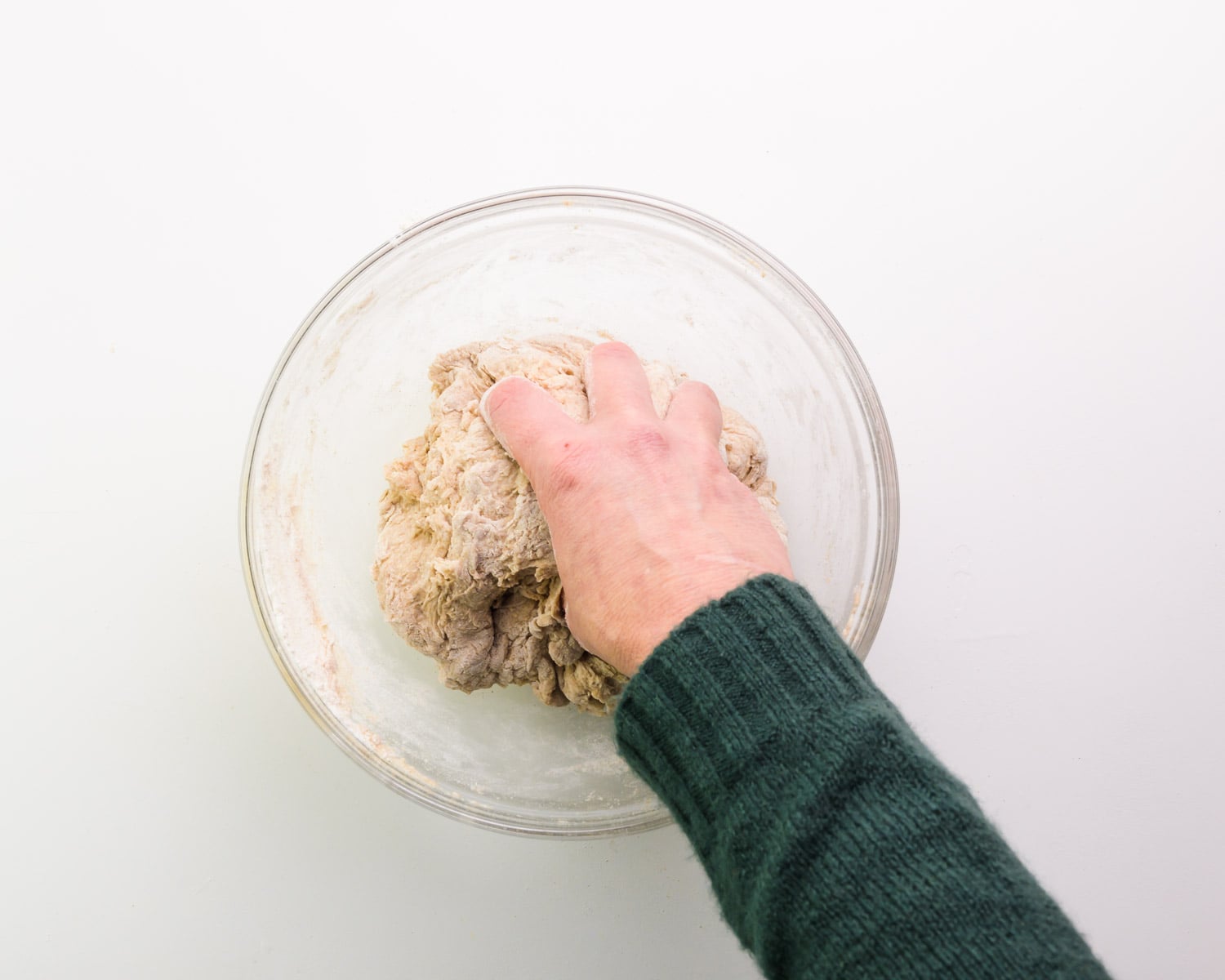 A hand is grabbing a ball of dough in a glass bowl.
