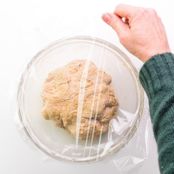 A hand is holding a piece of plastic wrap, covering a bowl of dough.
