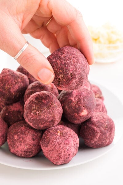 A hand holds a red velvet ball hovering over a stack of them on a plate. A bowl of white chocolate chips is in the background.