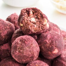 A red velvet ball has a bite taken out of it showing white chocolate chips inside. It sits on a stack of more of the energy balls.