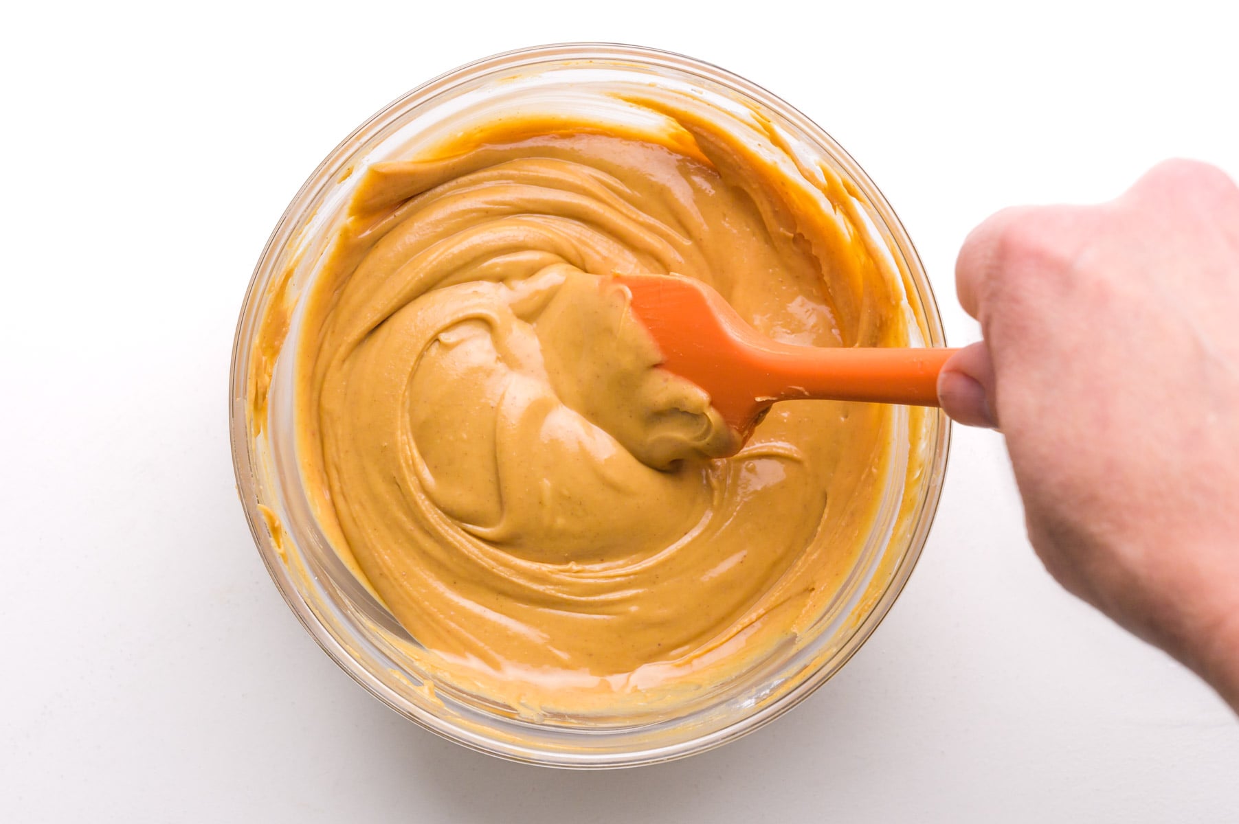 hand holds an orange spatula stirring peanut butter in a bowl.