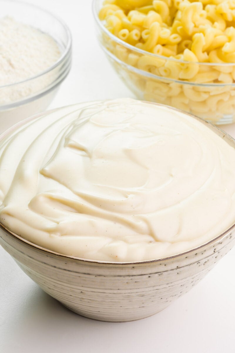 A bowl of vegan béchamel sauce since in front of a bowl of flour and bowl of cooked macaroni.