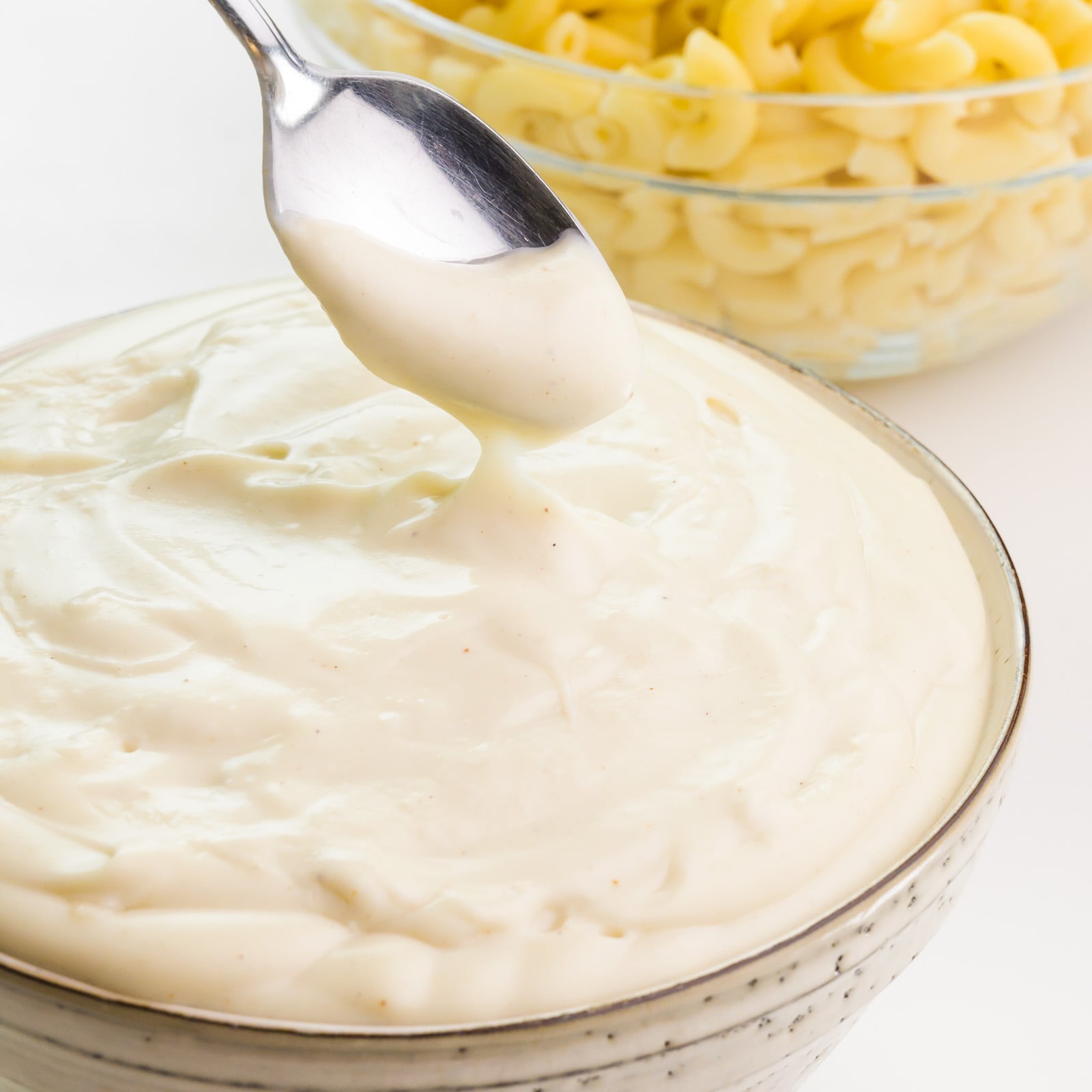 A spoon has some béchamel sauce in it and it is drizzling into the bowl. There's a bowl of macaroni in the background.
