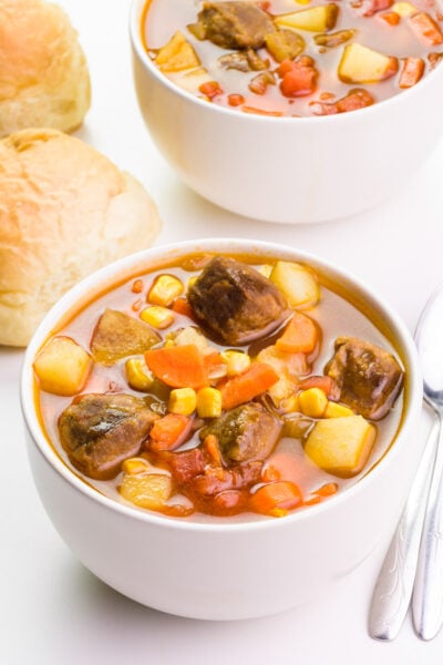 Looking down on a bowl of vegan beef stew with two spoons behind it, another bowl of the stew and dinner rolls are in the background.