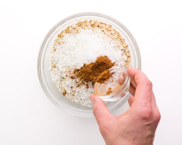 A hand holds a bowl of ground cinnamon, pouring it into a bowl with other ingredients like oats and coconut flakes.