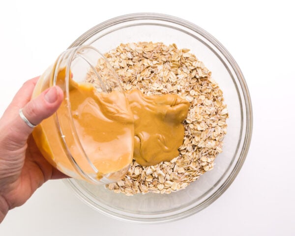 A hand holds a bowl of peanut butter, pouring it into a bowl with an oats mixture.
