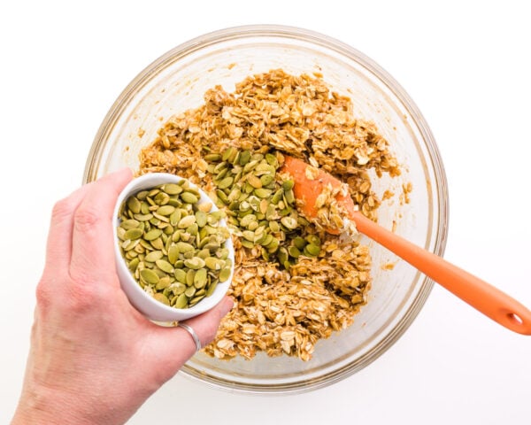 A hand holds a bowl of pumpkin seeds, pouring them into a bowl with cookie dough.