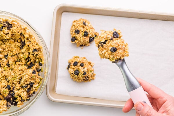 A hand holds a cookie dough scoop full of cookie dough over a baking pan with more cookie dough balls on it. There's a bowl with more dough on the side.