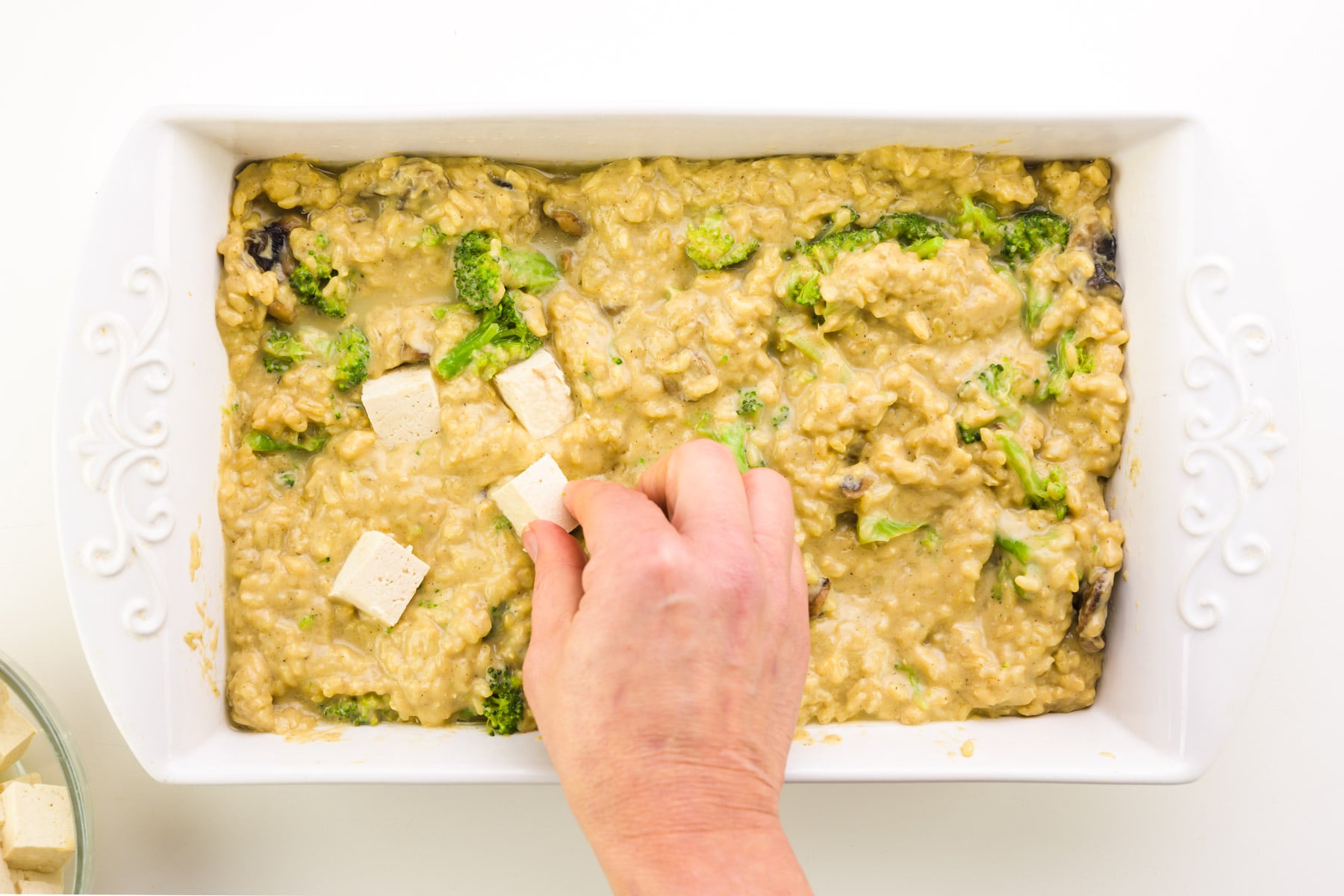 A hand presses tofu cubes into a casserole dish.