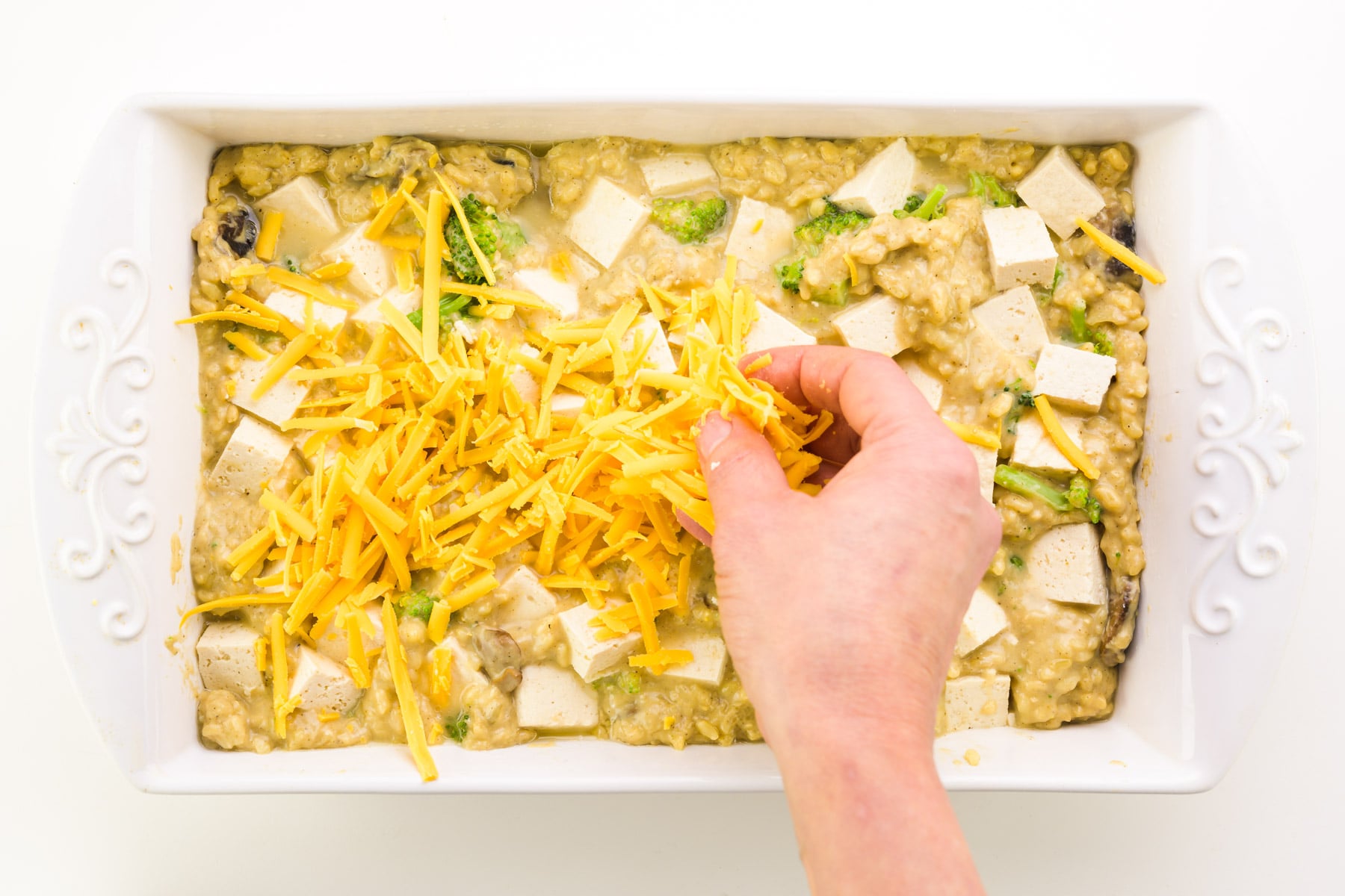 A hand spread shredded cheddar cheese over a tofu casserole.