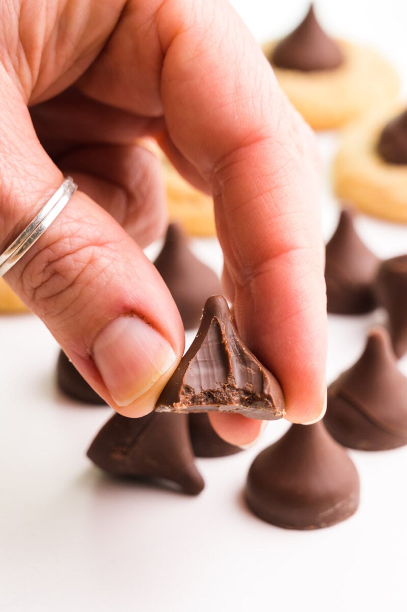 A hand holds a vegan chocolate kiss with a bite taken out. There are more of the candies and cookies in the background.