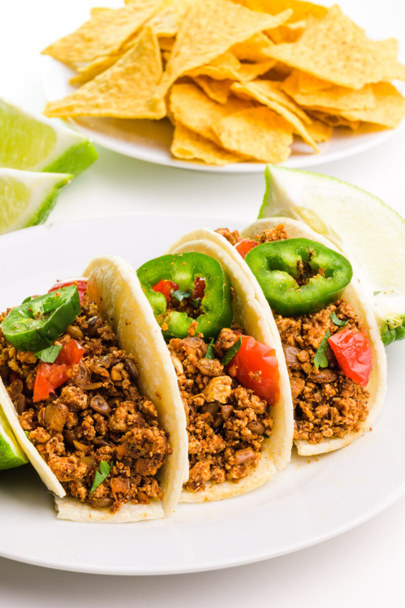Three soft tacos sit on a plate, topped with vegan chorizo and jalapeños. There are lime slices and tortilla chips in the background.