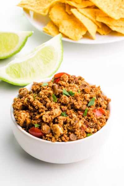 A bowl of vegan chorizo sits in front of lime slices and a plate with tortilla chips.