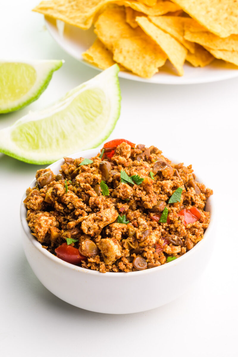 A bowl of vegan chorizo sits in front of lime slices and a plate with tortilla chips.