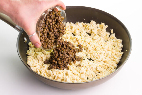 A hand holds a bowl of cooked lentls, pouring them into a skillet with tofu pieces.