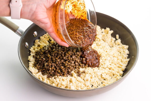 A hand is pouring a bowl of sauce into a skillet with tofu and lentils.