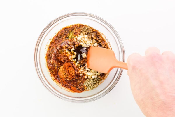 A hand holds a spatula stirring together sauce ingredients in a bowl.