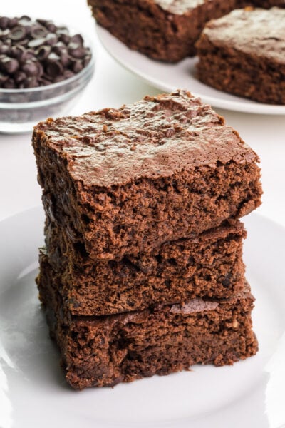 A stack of three gluten-free brownies sit on a plate in front of a bowl of chocolate chips ad more brownies in the background.
