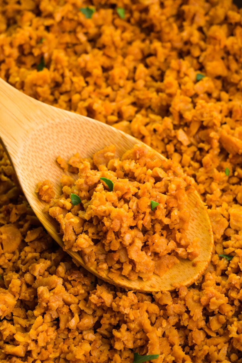 A wooden spoon holds vegan ground beef in a skillet with more of the crumbles around it.