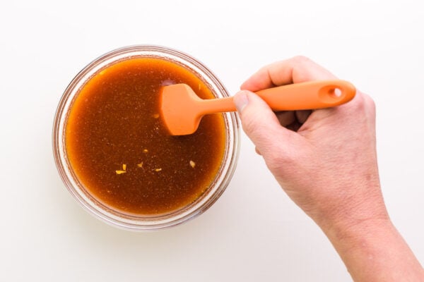 A hand holds a rubber spatula, stirring sauce in a bowl.