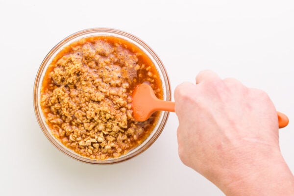 A hand holds a rubber spatula, stirring a bowl with sauce and textured vegetable protein.